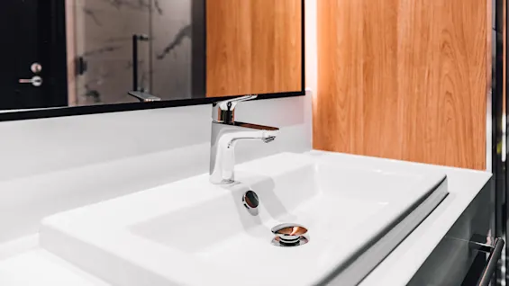 Chrome faucet stands over a white ceramic sink, reflecting wood-paneled walls and a bathroom mirror. A partially visible shower area is in the background.