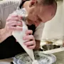 A chef concentrates while using a piping bag to shape cream onto a decorative plate in a professional kitchen.