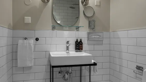 A clean and modern bathroom with a white sink, an oval mirror, and bright wall sconces. White subway tiles line the walls.