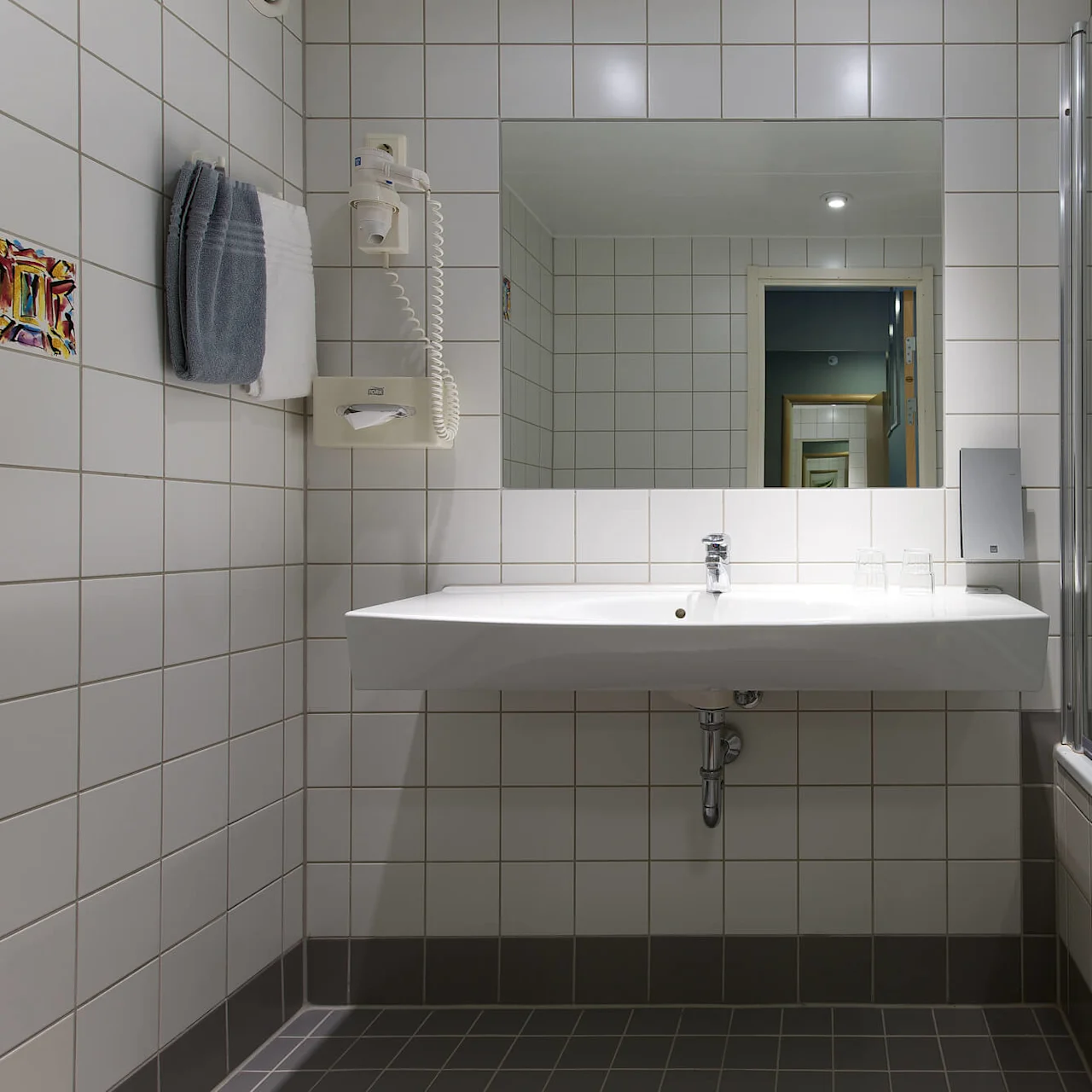 A white sink is mounted on a tiled bathroom wall, with a mirror above. A towel, hairdryer, and glass are nearby. A bathtub with a transparent screen is on the right.