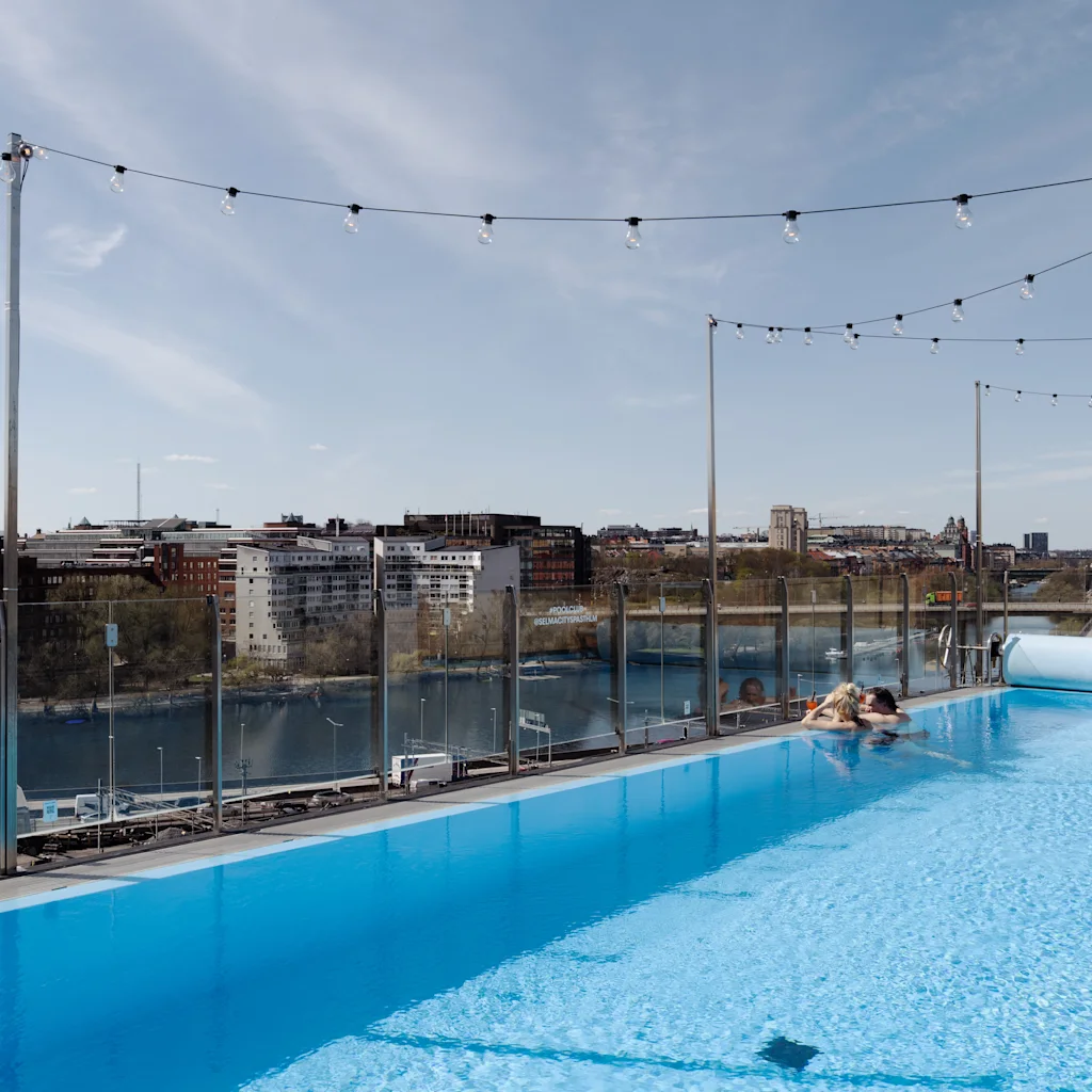 Takpool med en person som kopplar av vid kanten, omgiven av ljusslingor. Utsikt över en stadsbild med byggnader och en flod under klar himmel.