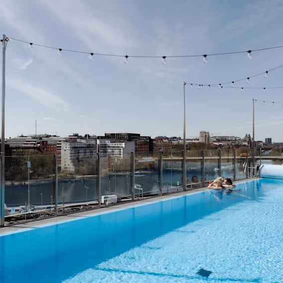 Takpool med en person som kopplar av vid kanten, omgiven av ljusslingor. Utsikt över en stadsbild med byggnader och en flod under klar himmel.