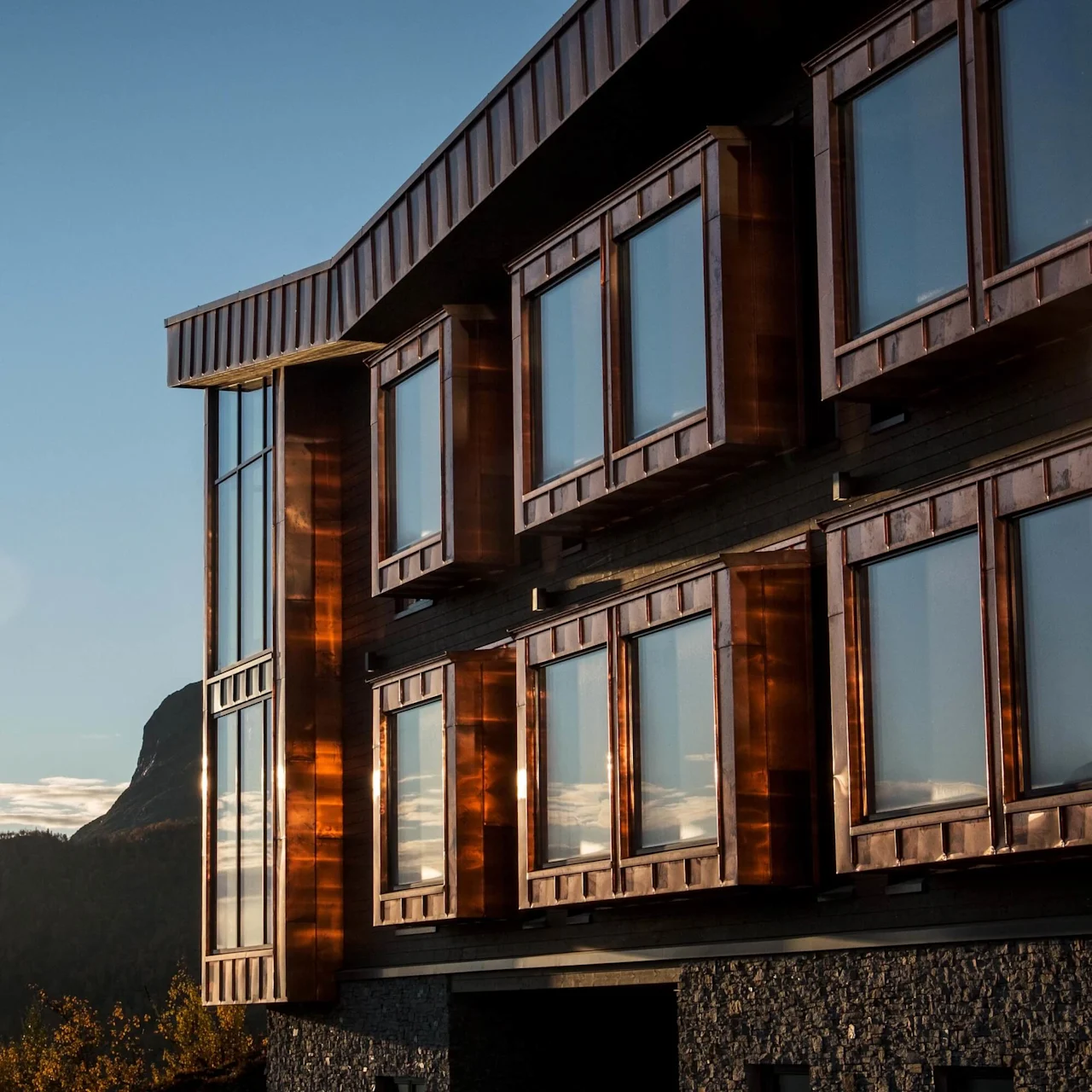 A modern building with large reflective windows, partially bathed in sunlight, stands against a backdrop of distant hills and a clear blue sky.