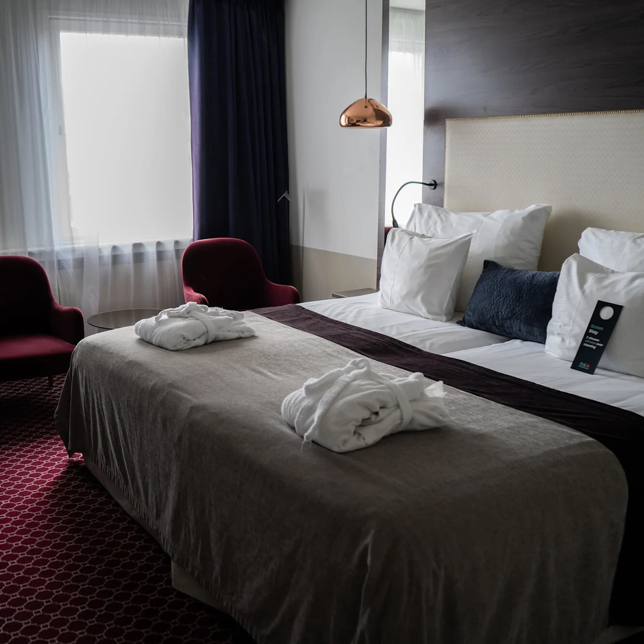 En prydligt bäddad hotellsäng med morgonrockar och en skylt på kuddarna, omgiven av två röda stolar, ett bord och stora ljusa fönster i ett mysigt rum.