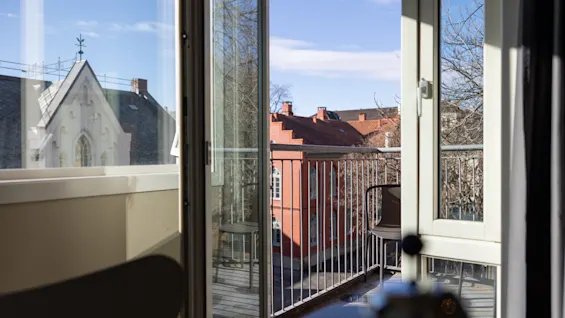 Utsikt från ett rum mot en balkong med en stol, med utsikt över byggnader med röda tak och kala träd under en blå himmel.