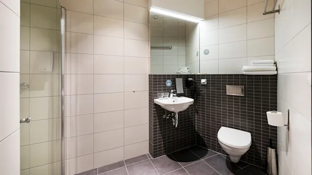 A modern bathroom features a wall-mounted sink and toilet in a tiled room. A glass shower enclosure is on the left, with folded towels on a shelf.