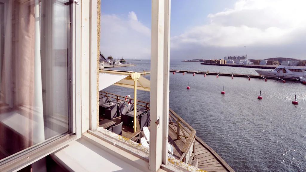 Outdoor terrace by the water, with chairs and tables under an umbrella, viewed through an open window. A yacht and buoys are visible on the calm sea.