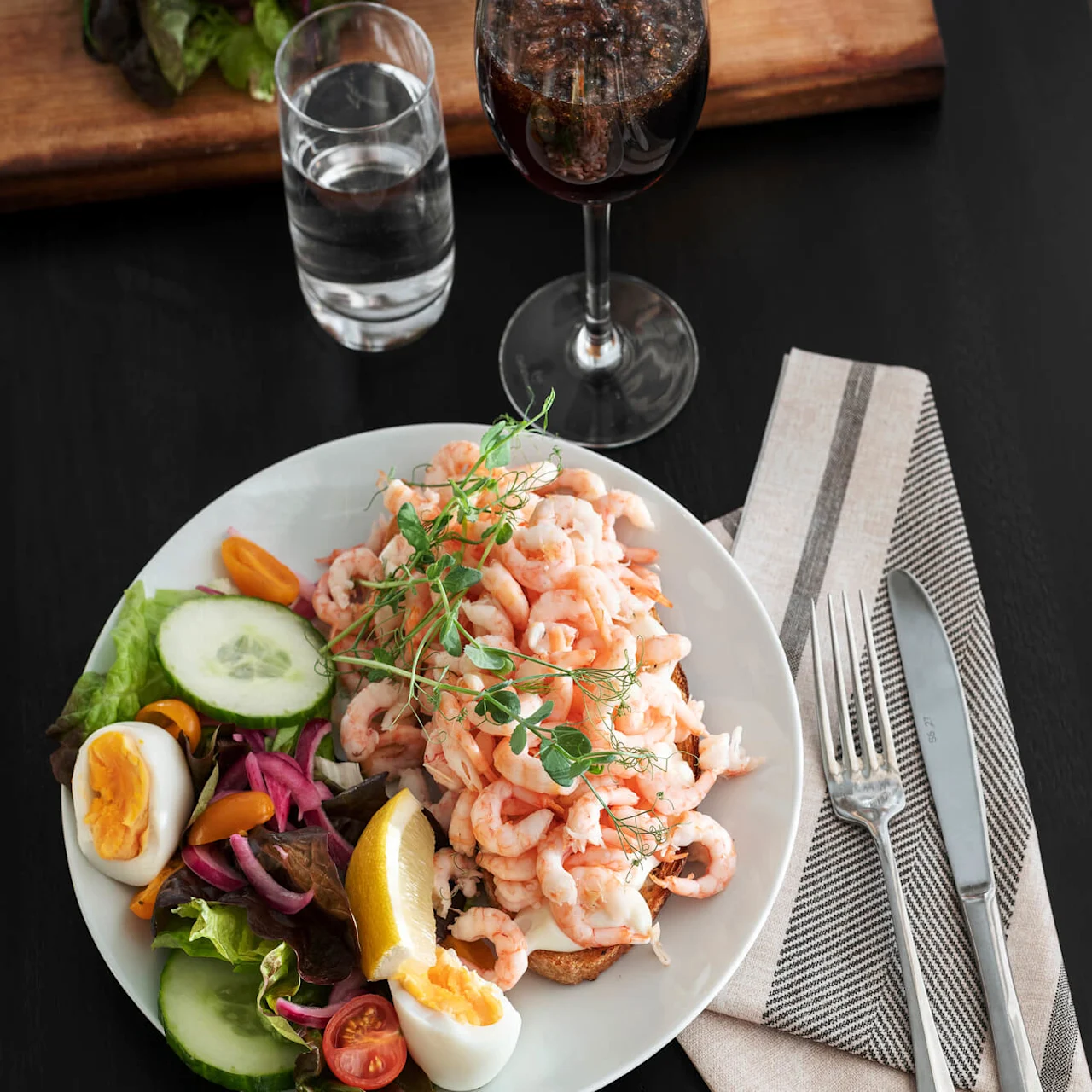 Tallerken med rekesalat, agurk, kokt egg, tomater og grønne blader på et svart bord. Et glass vann, vin og bestikk ligger ved siden av på en stripet serviett.