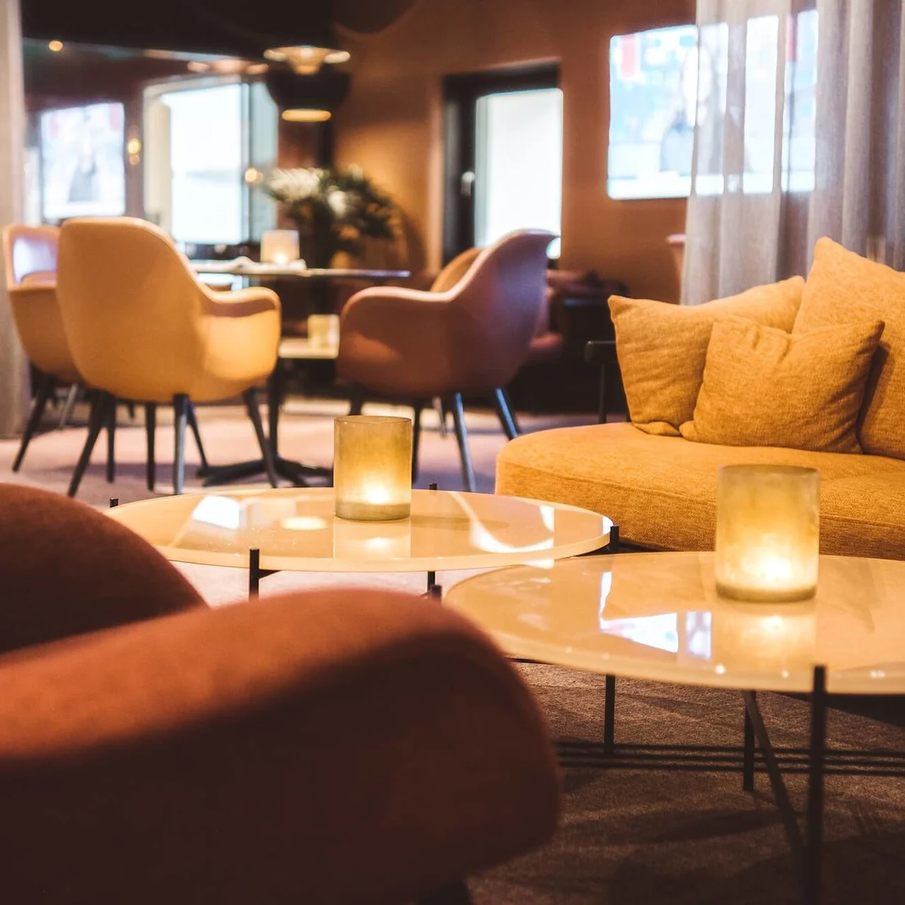 En hyggelig lounge med beige sofaer og stole omkring lave, runde borde med uoplyste lys, placeret i et varmt oplyst, elegant rum.