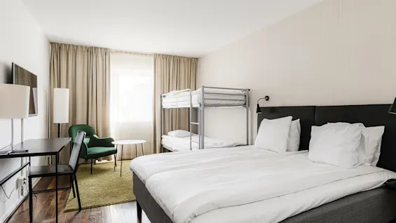 Ett hotellrum med dubbelsäng och våningssäng, bredvid en grön fåtölj och ett litet bord; ett skrivbord med stol finns i närheten, allt under mjukt ljus.