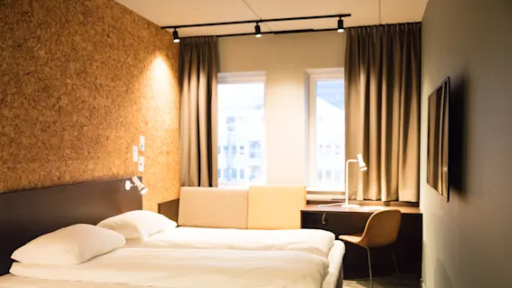 A double bed is positioned against a cork-textured wall, with bedside lamps. Adjacent, a desk with a chair and lamp sits by two curtained windows, in a minimalist hotel room.