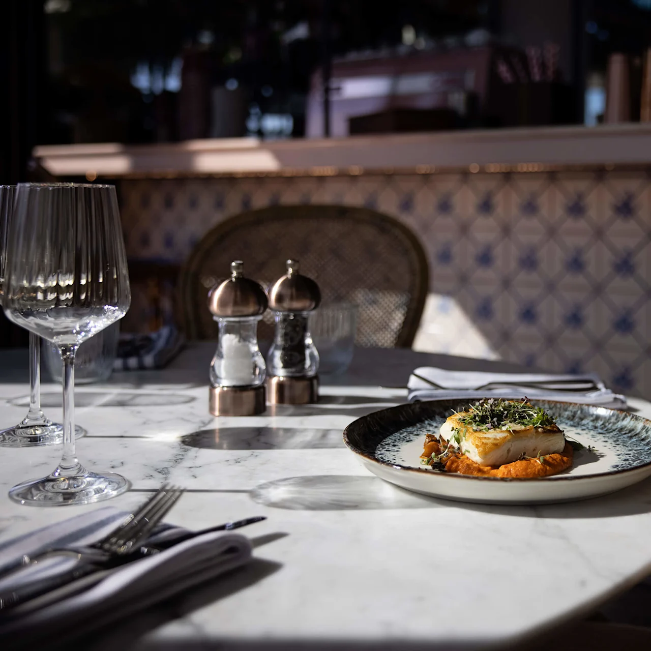 Plate with a gourmet dish sits on a marble table. Sunlight highlights the setting, including glassware and a patterned tiled wall, in an elegant dining area. 