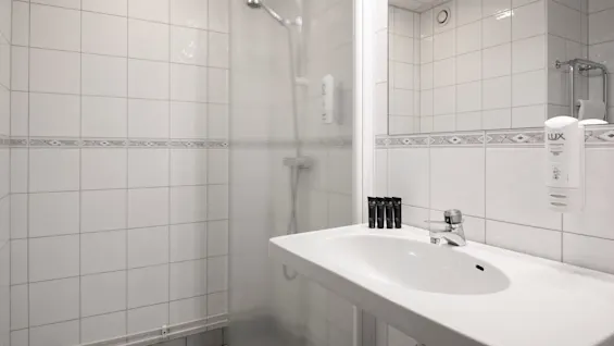 En vit vask med toalettartiklar står bredvid en dusch av frostat glas i ett kaklat badrum. Text på dispenser: "LUX."