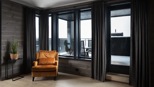 Orange armchair sits in a modern room with large windows, dark curtains, and wooden floors. A small table with a potted plant decorates the corner, overlooking a balcony.