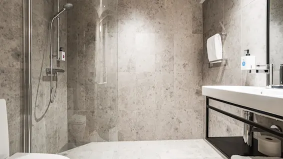 A modern bathroom with light grey tiled walls and floor, featuring a walk-in shower with a glass partition and a white sink with a mirror.