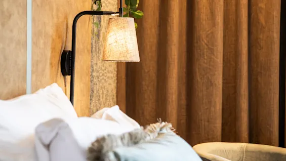 Säng med fluffiga kuddar och mjuk sänggavel, placerad bredvid en väggmonterad lampa. En grön växt hänger ovanför, med bruna gardiner och en fåtölj i bakgrunden.