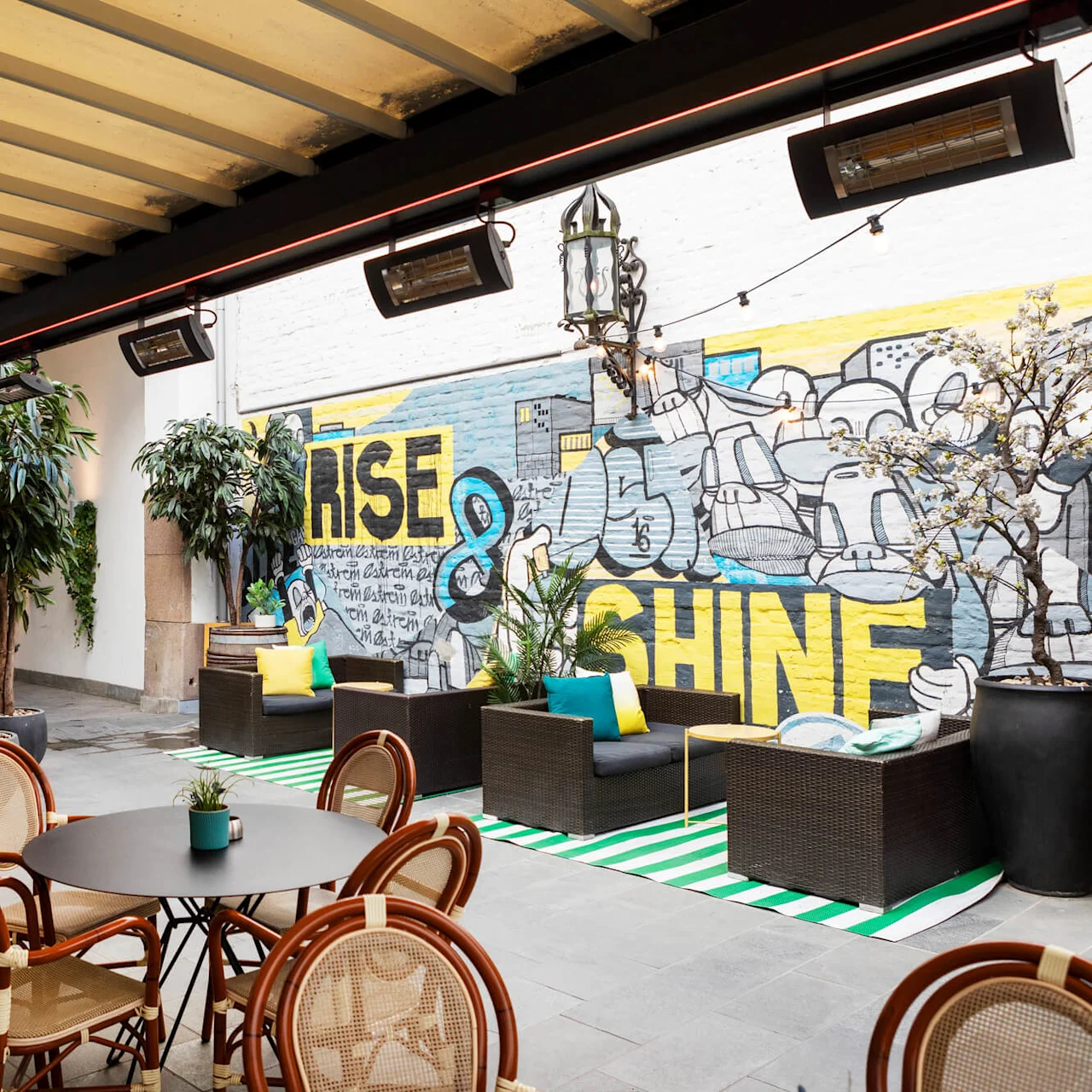 Uteområde med runde bord og rottingstoler under en pergola. Et veggmaleri har teksten "RISE & SHINE", omgitt av planter i potter og puter på sofaer.