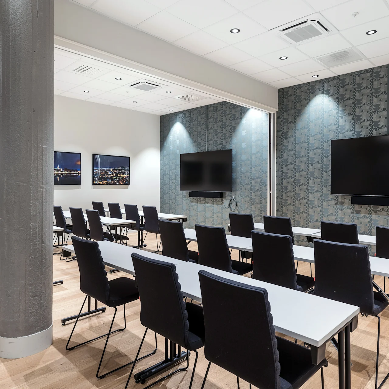 Rivistöt mustia tuoleja ovat käännettyjä seinällä oleville näytöille konferenssihuoneessa, jossa moderni ilme korostuu koristeellisella tapetilla ja puulattialla.