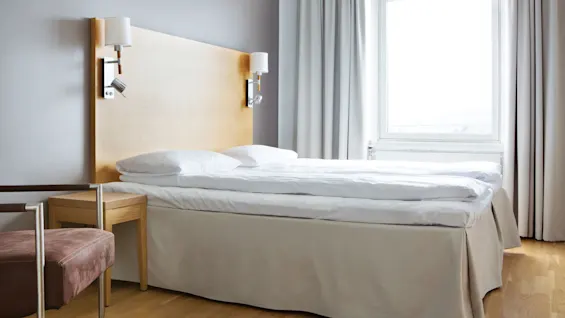 En seng med hvitt sengetøy står ved siden av et stort vindu i et lyst rom. Et nattbord i tre og en polstret stol gir komfort.