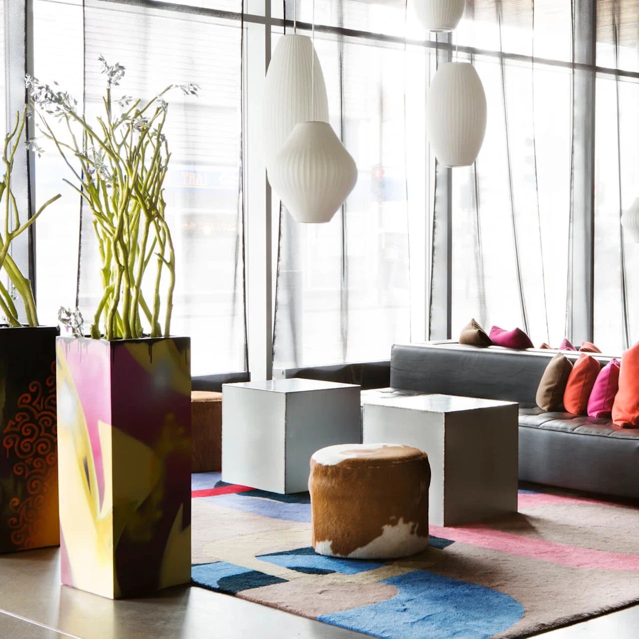 Sofa med fargerike puter, står stille, i et moderne rom med store vinduer, livlige planter, geometriske bord og hengende hvite lamper.