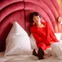 A person in a red outfit sits on a bed against a plush, curved headboard. White pillows surround them. A bedside lamp glows warmly beside the bed.