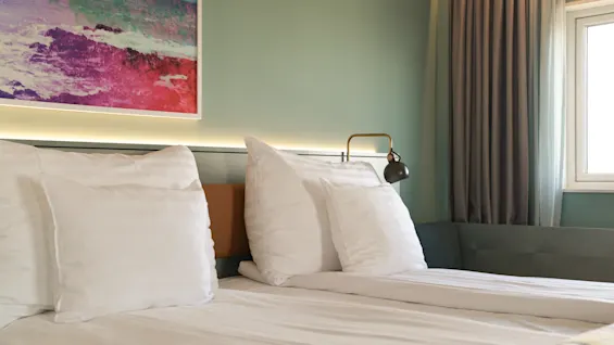 En pænt redt dobbeltseng med hvide puder og lagner står i et blødt oplyst rum, flankeret af en moderne lampe. Et farverigt abstrakt maleri hænger over hovedgærdet.