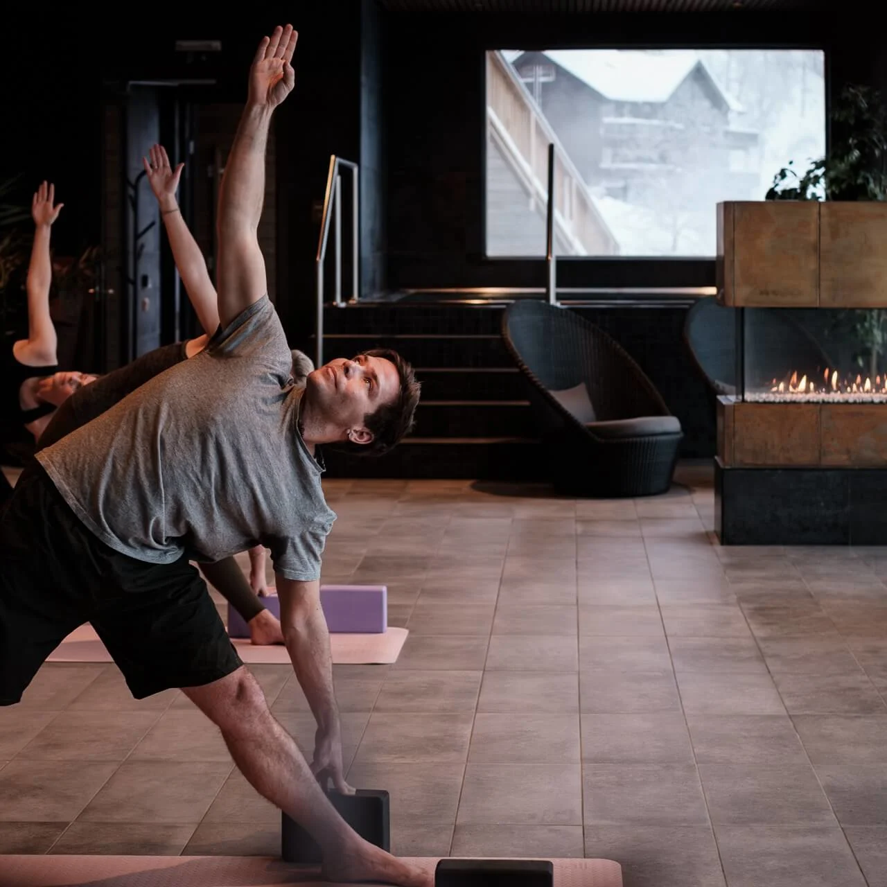 People practice yoga poses in a modern room with a tiled floor and a lit fireplace. Large window shows a snowy exterior.