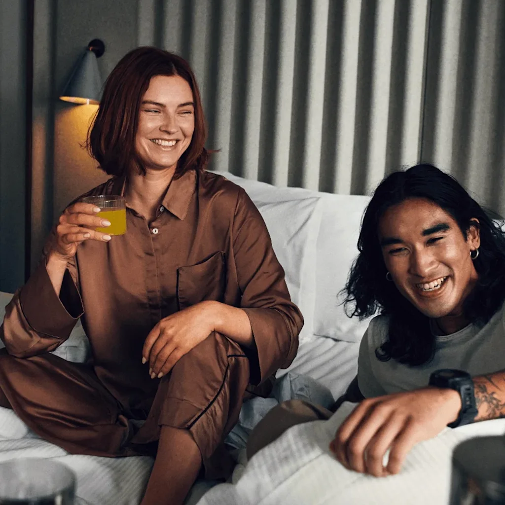 A woman in brown pajamas holds a glass of orange juice, smiling beside a man on a bed in a cozy room with dim lighting and gray curtains.