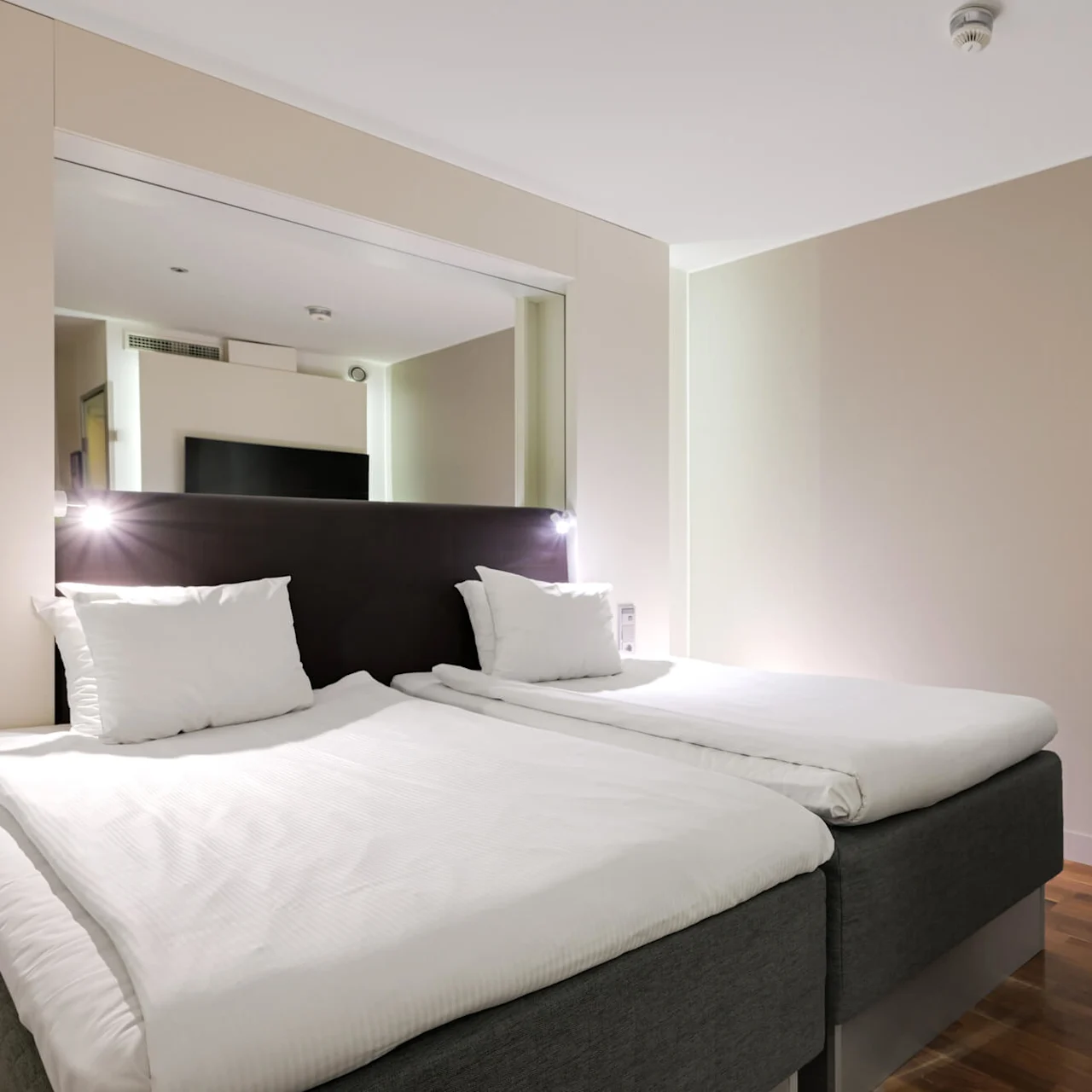 Två prydligt bäddade enkelsängar står med en sänggavel, upplysta av läslampor, i ett modernt, minimalistiskt hotellrum med trägolv och en väggmonterad TV.