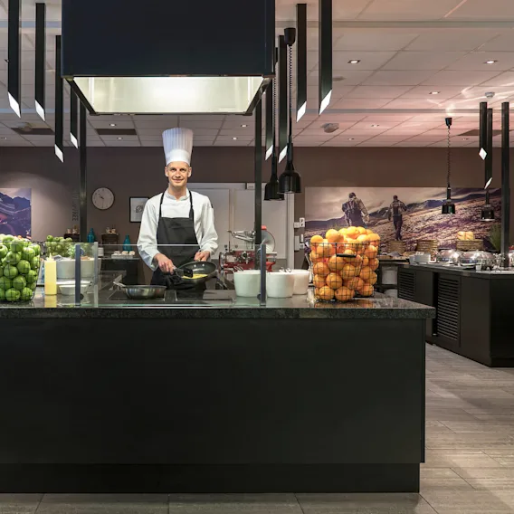 A chef stands smiling, cooking in a modern kitchen with fruit baskets and buffet stations, under sleek pendant lights, surrounded by wall art depicting outdoor scenes.