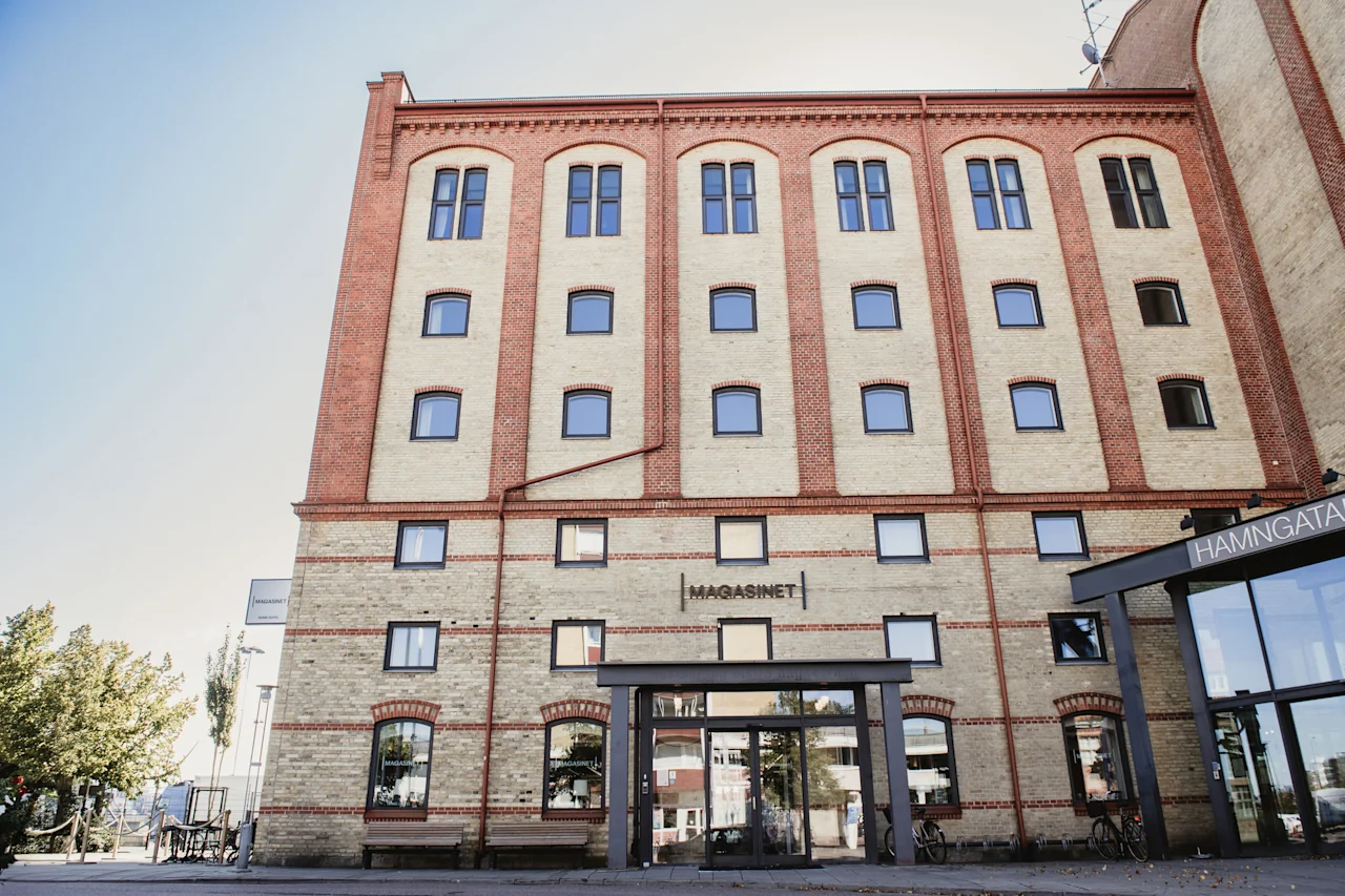 Suuri tiilirakennus, jossa on paljon ikkunoita ja moderni lasisisäänkäynti. Tekstit 'MAGASINET' ja 'HAMNGATAN' näkyvät.