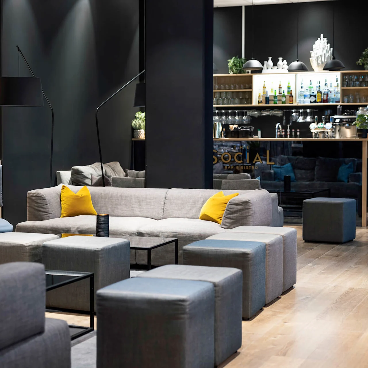 Grå sofaer med gule puter og kubestoler i en moderne lounge setting med mørke vegger og en bar. Tekst: "SOCIAL BAR & BISTRO."