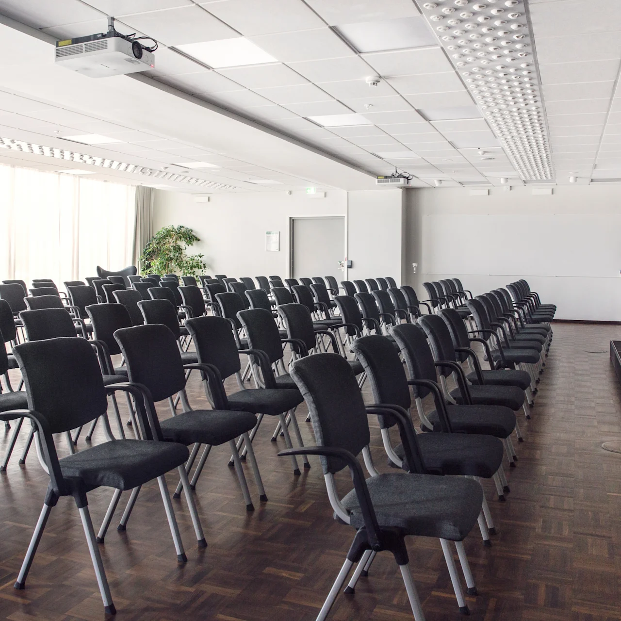 Rader av tomma svarta stolar står vända mot en liten scen med ett podium och stolar i ett konferensrum, upplyst av naturligt ljus från stora fönster till vänster.