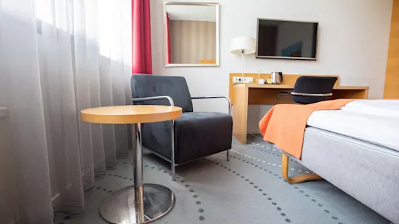 Et moderne hotellrom viser en svart lenestol ved siden av et lite trebord, med en skrivebord og TV i bakgrunnen. Røde gardiner rammer inn vinduet.