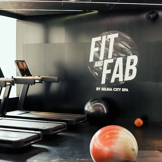 Löpband står oanvända i ett gym. En färgglad träningsboll och vikter ligger på det mörka golvet. "FIT AND FAB BY SELMA CITY SPA" visas på väggen.