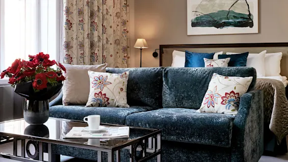 En blå sofa med blomstrede puder står i en hyggelig stue. Et sofabord har en kop og aviser. Røde blomster pryder en vase nær mønstrede gardiner.