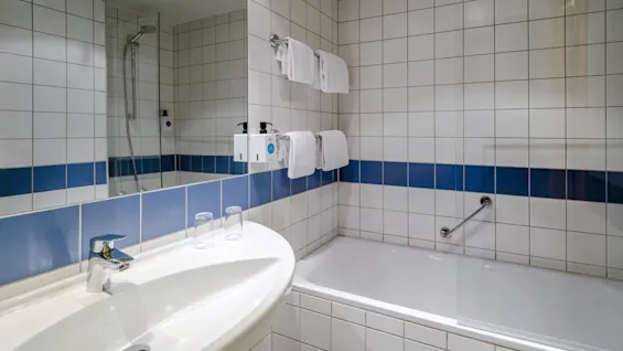 Håndvask med hane og to glas, ved siden af et badekar med brusehoved og sæbedispensere, i et flisebelagt badeværelse med en blå stribe og pænt hængte hvide håndklæder.