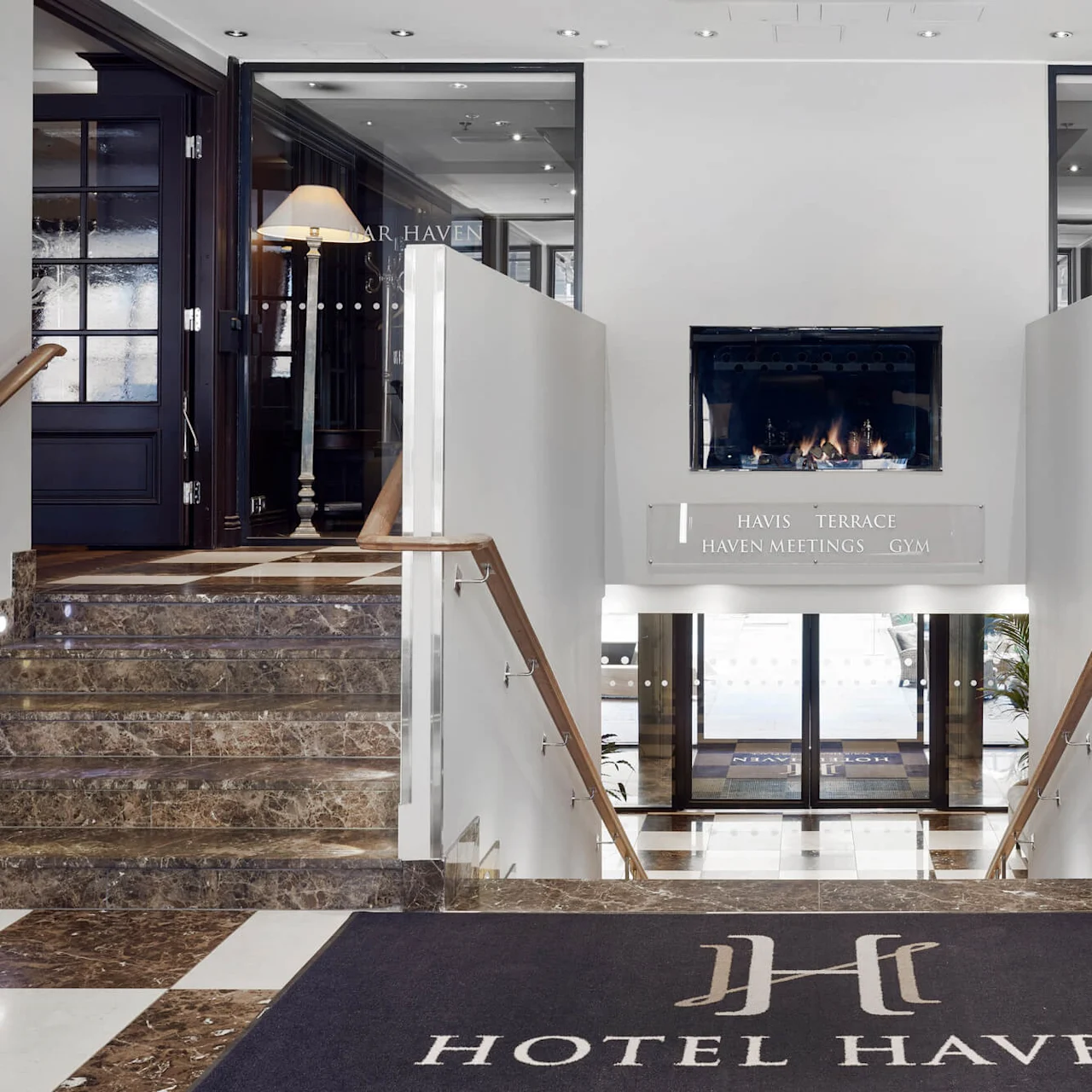 Entrance hall with marble stairs descending to glass doors. A fireplace is centered above, flanked by decorative lamps. Sign reads "Havis Terrace Haven Meetings Gym" and mat says "Hotel Haven."