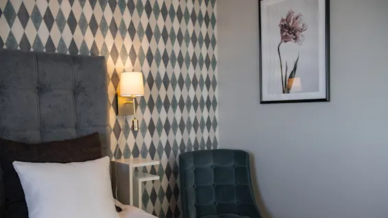 Bed in a bedroom, featuring a gray upholstered headboard and white pillow, beside a small nightstand and lamp. A blue chair and framed flower artwork decorate the patterned wall.