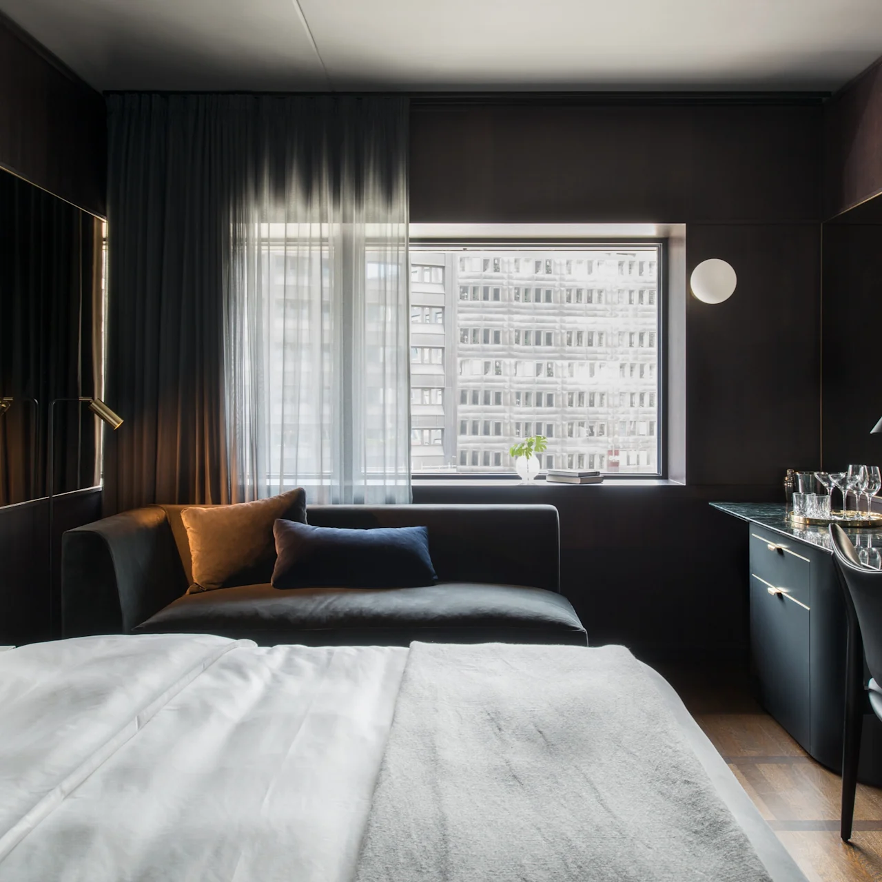 Et moderne hotellrom har en pent oppredd seng, en mørk sofa med puter og et skrivebord med stol. Et stort vindu viser en byutsikt utenfor.