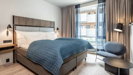 En pænt redt seng med blåt tæppe står mod et træhovedgærde, flankeret af væglamper. En moderne stol og bord ved vinduet, indrammet af gardiner. Hyggelig stemning.