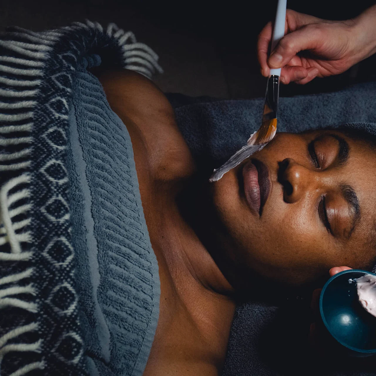 En person ligger på en massagebänk med slutna ögon, medan en annan applicerar en ansiktsmask med en borste. De är inlindade i en handduk, vilket antyder en behandling på ett spa.