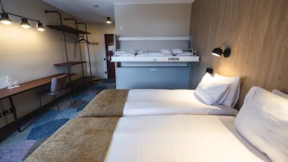 A hotel room with a double bed, neatly made, has a bunk bed on the wall. Modern lighting fixtures highlight a desk and chair along a patterned carpet floor.