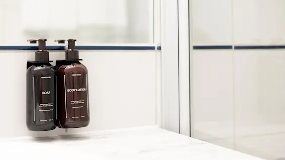 Kaksi annostelijapulloa, toinen SAIPPUA ja toinen BODY LOTION, valkoisella kaakeloidulla kylpyhuoneen seinällä.