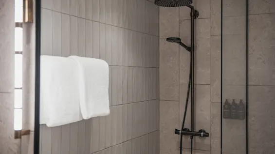 A modern hotel bathroom shower with dark fixtures, white towels, and tiled walls at Home Hotel Tapetfabriken.