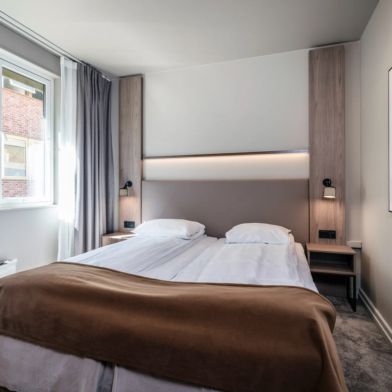 A neatly made double bed with white linens and a brown blanket, flanked by wooden side panels with lamps, in a bright room with a large window and framed wall art.