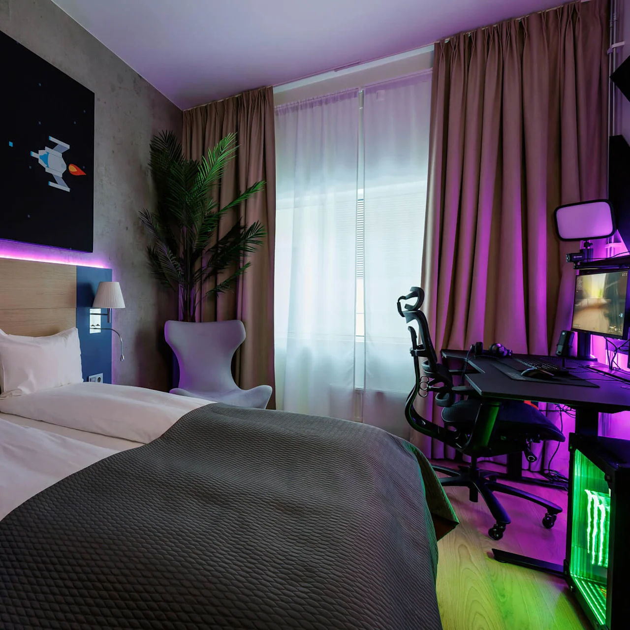 A bed with white sheets is illuminated by purple LED lights beneath a space-themed picture. Nearby, a gaming setup with a chair and computer is visible, framed by curtains.