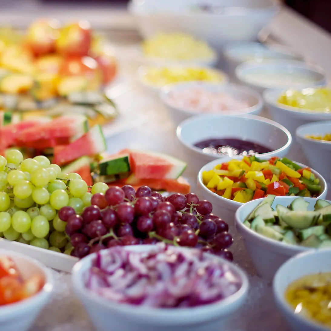 Et udvalg af frisk frugt og grøntsager, herunder druer, vandmelon og peberfrugter, er præsenteret i skåle og tallerkener på et bord og skaber en farverig buffetanretning.