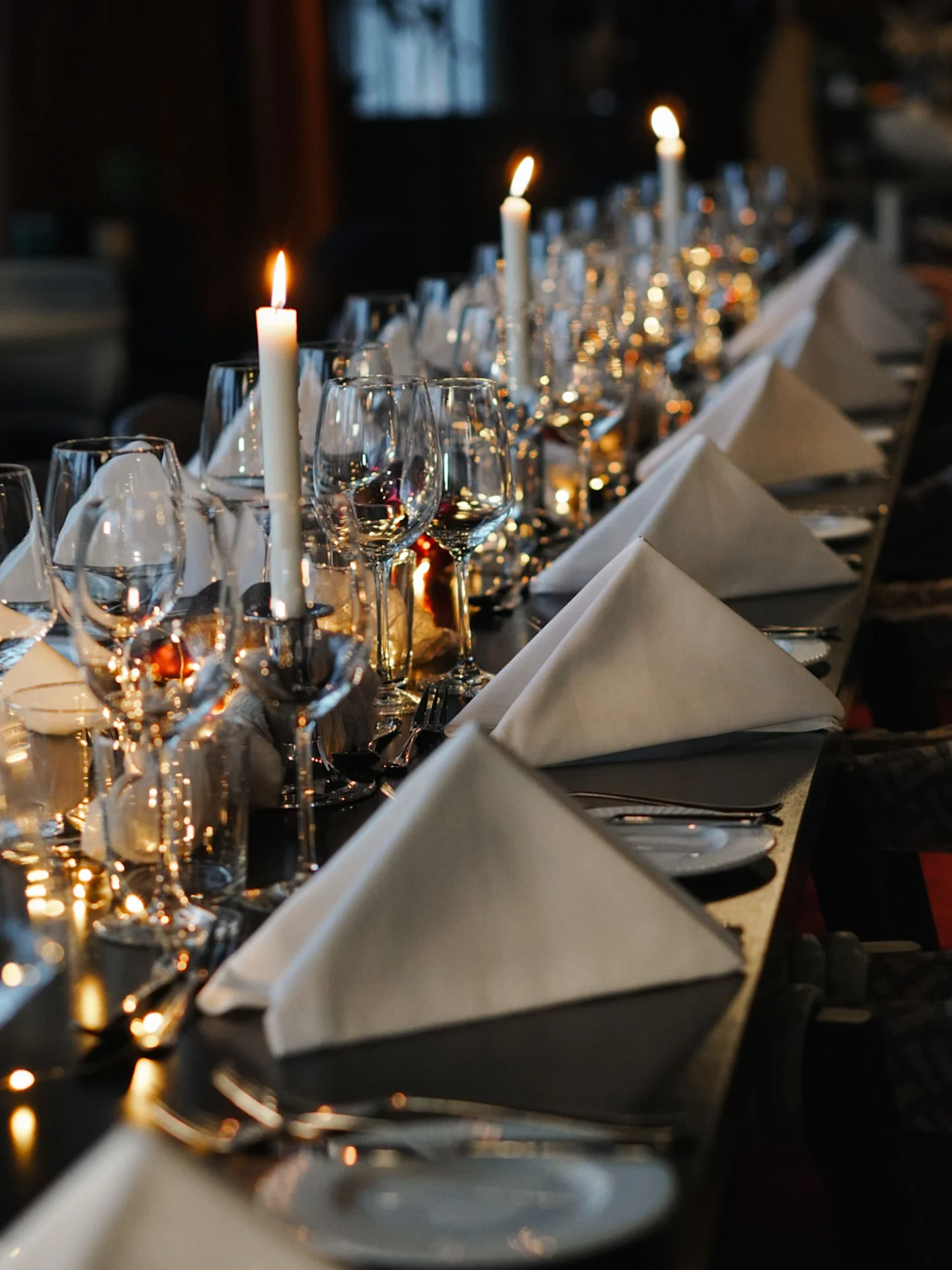 Elegant spisebord dekket med hvite lys, brettede servietter og glass; svakt opplyst, og skaper en formell, intim atmosfære.