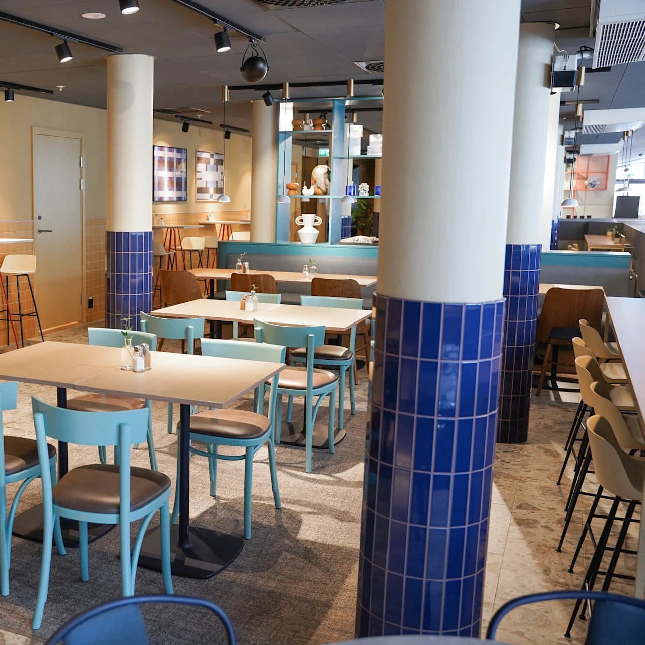 A bright café with turquoise chairs around wooden tables. Blue-tiled columns and potted plants enhance the aesthetic. Bar stools line the back wall, creating a cozy atmosphere.